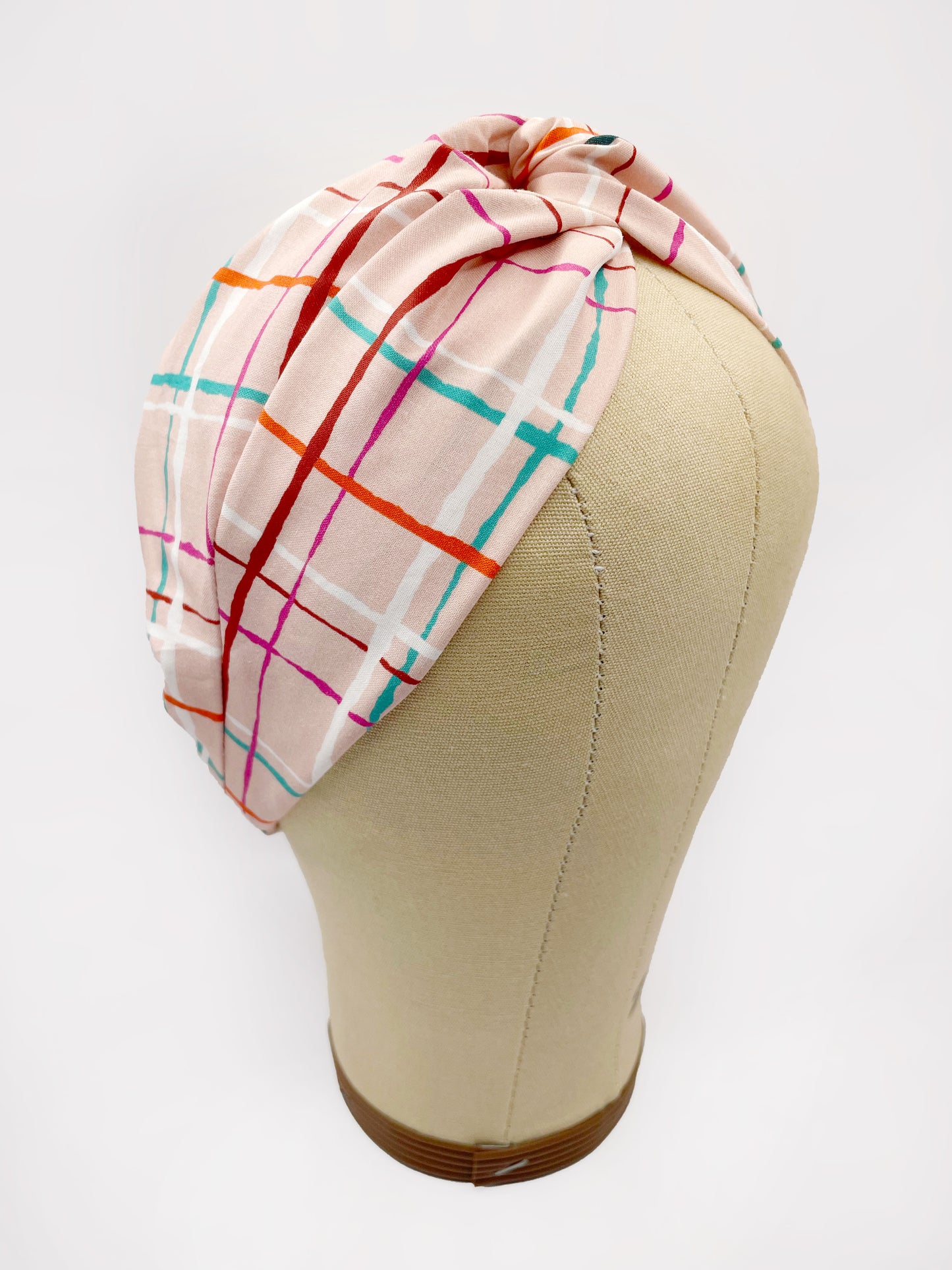 A fabric headband featuring a colorful and modern plaid design with vibrant shades of pink, red, teal, and orange on a soft pastel background. The headband is shown on a mannequin head.