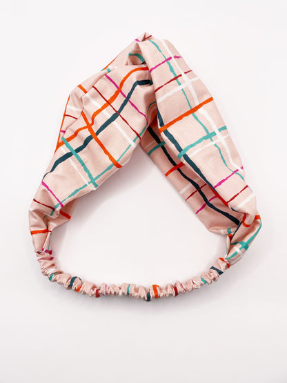 A fabric headband featuring a colorful and modern plaid design with vibrant shades of pink, red, teal, and orange on a soft pastel background. The headband has an elastic band in the back.