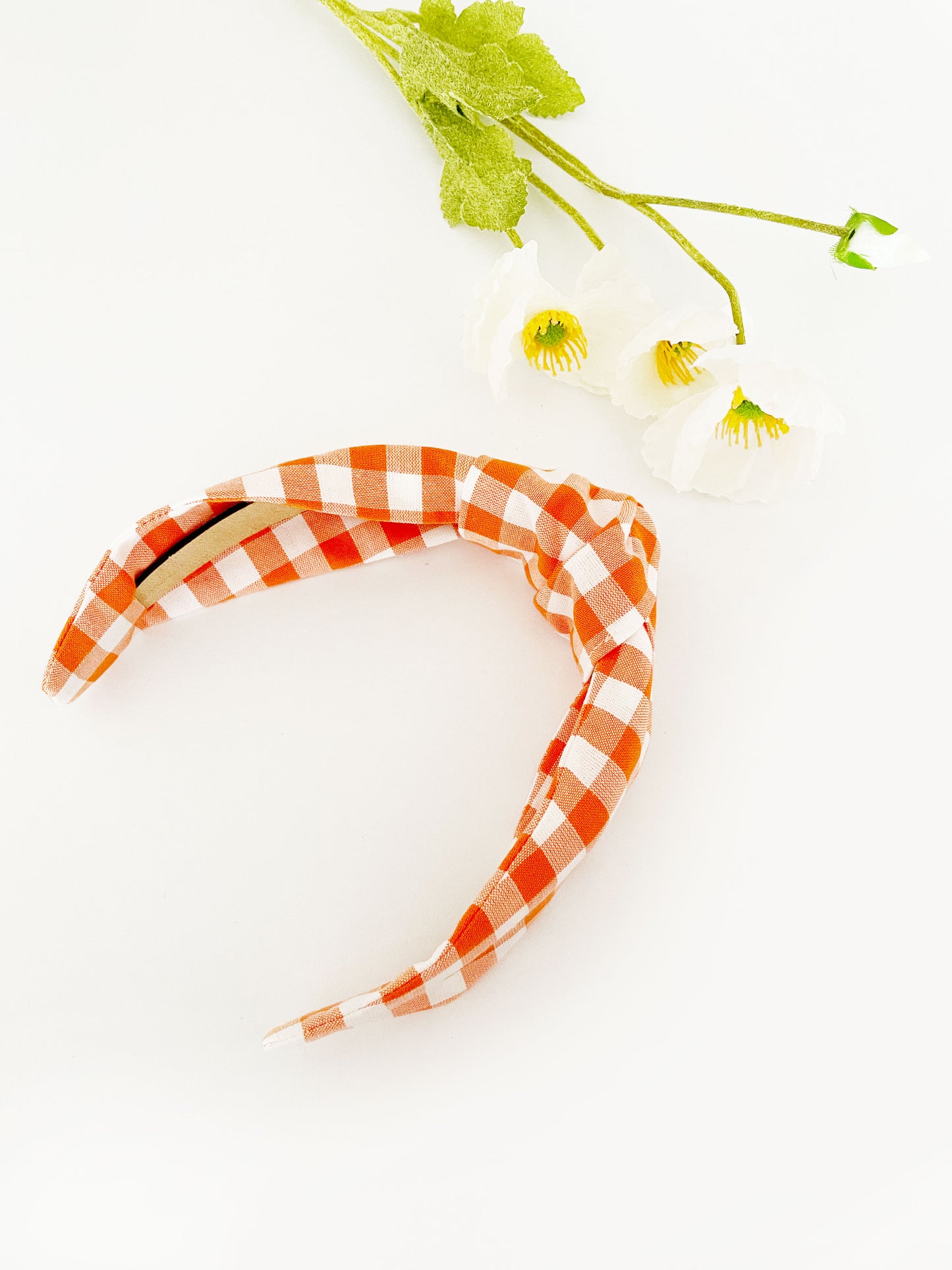 A knotted headband with an orange and white gingham plaid print. The headband is next to white artificial flowers.