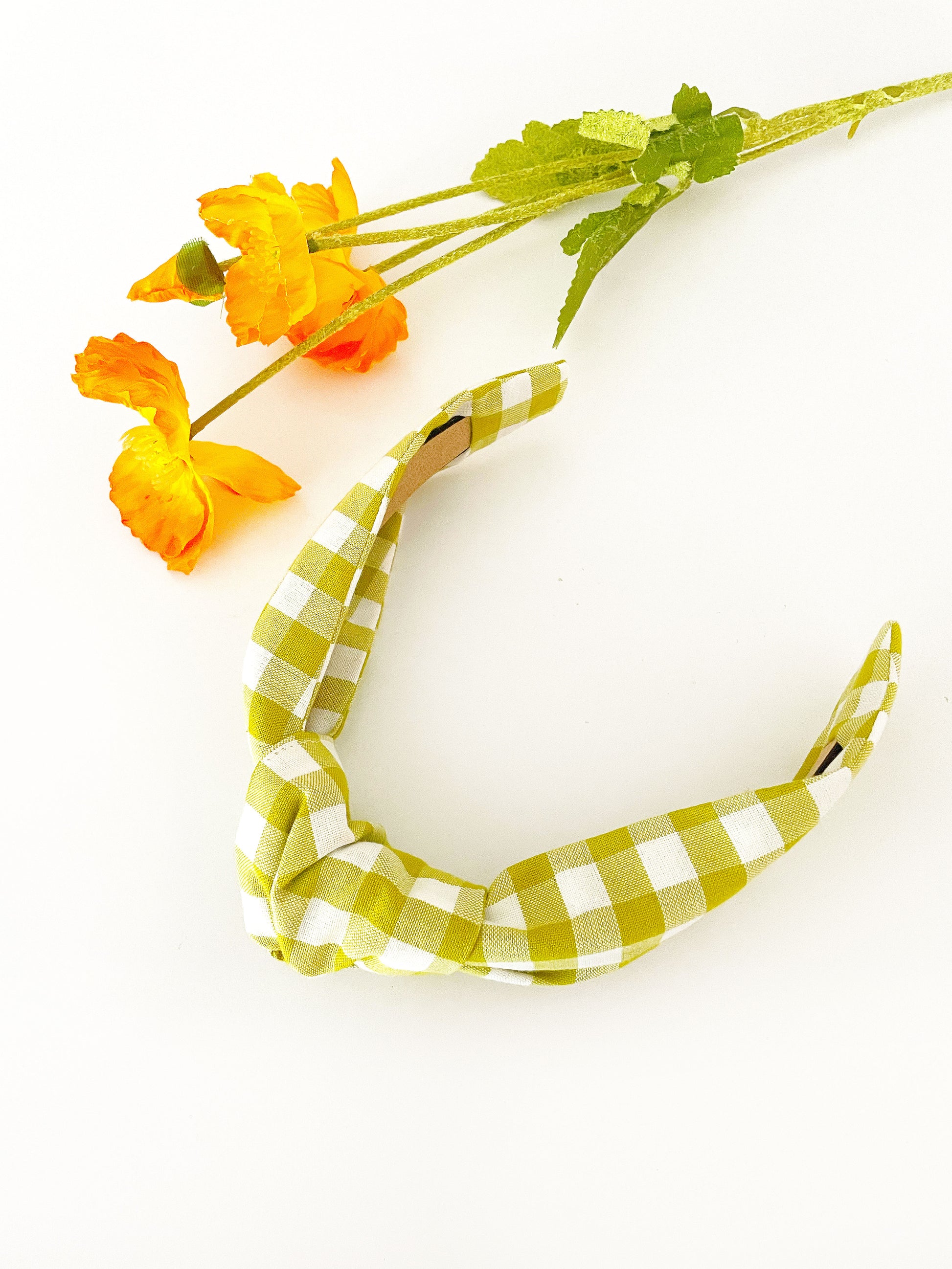 A knotted headband with a green and white gingham plaid print. The headband is next to orange artificial flowers.