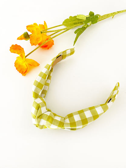 A knotted headband with a green and white gingham plaid print. The headband is next to orange artificial flowers.