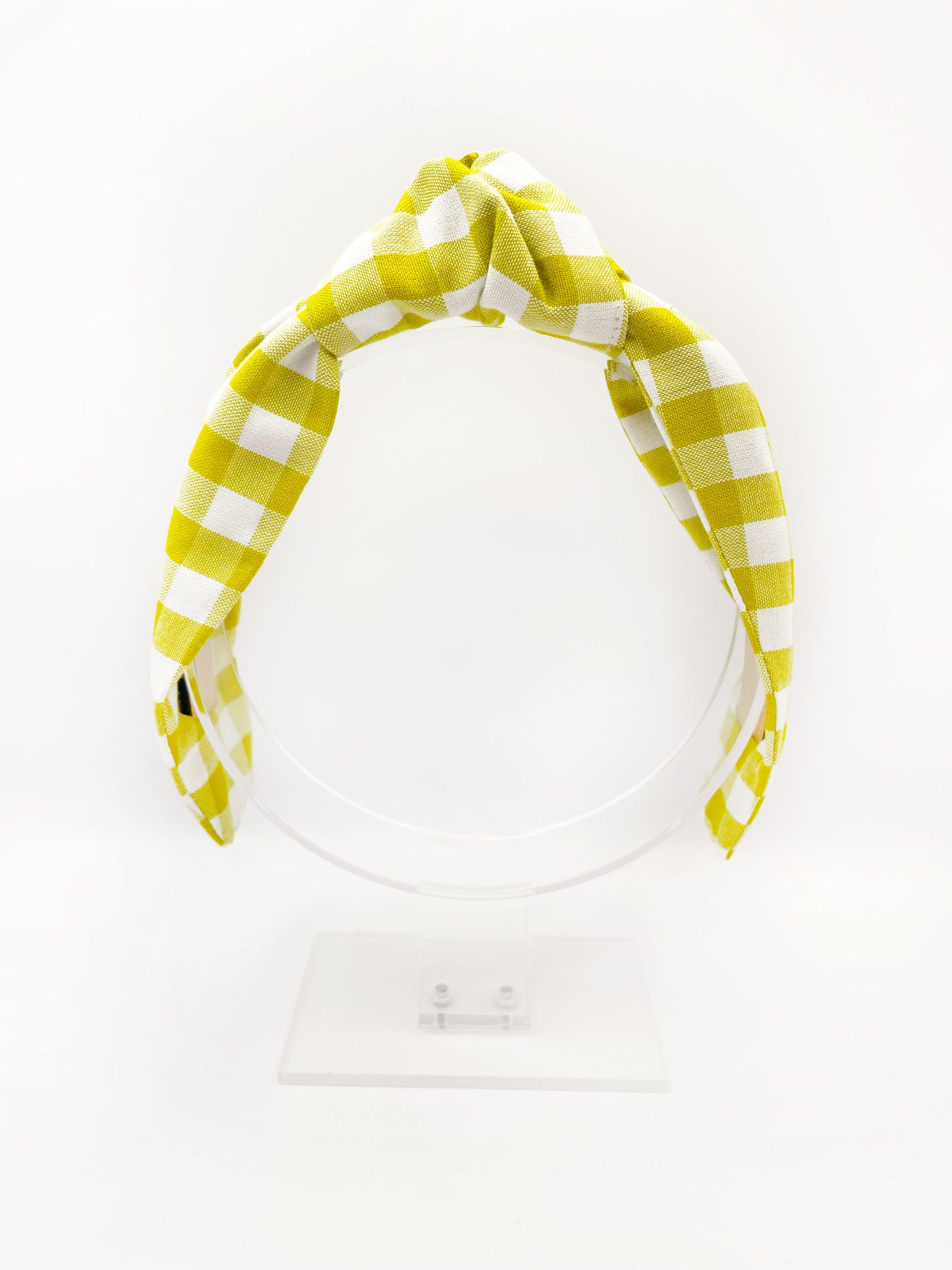 A knotted headband with a green and white gingham plaid print. The headband is modeled on an acrylic display stand.