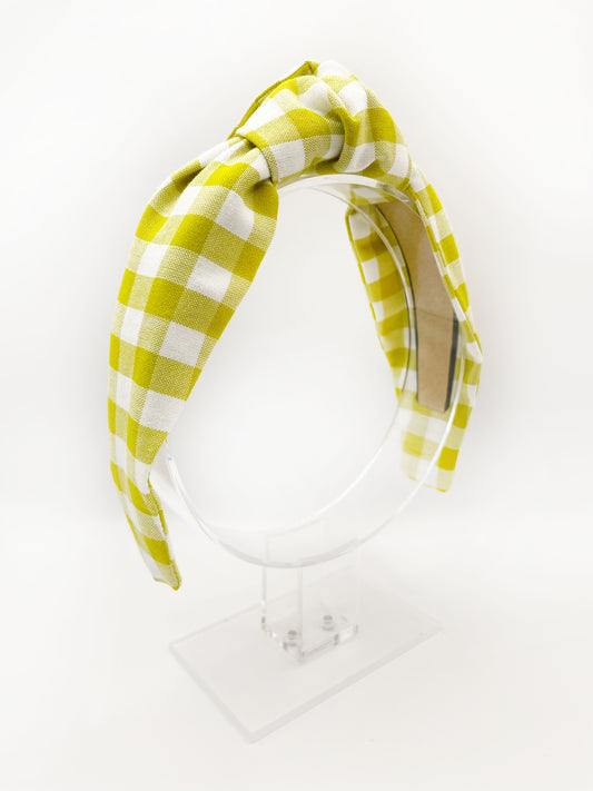 A knotted headband with a green and white gingham plaid print. The headband is modeled on an acrylic display stand.