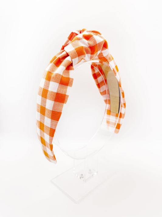 A knotted headband with an orange and white gingham plaid print. The headband is displayed on an acrylic headband display.