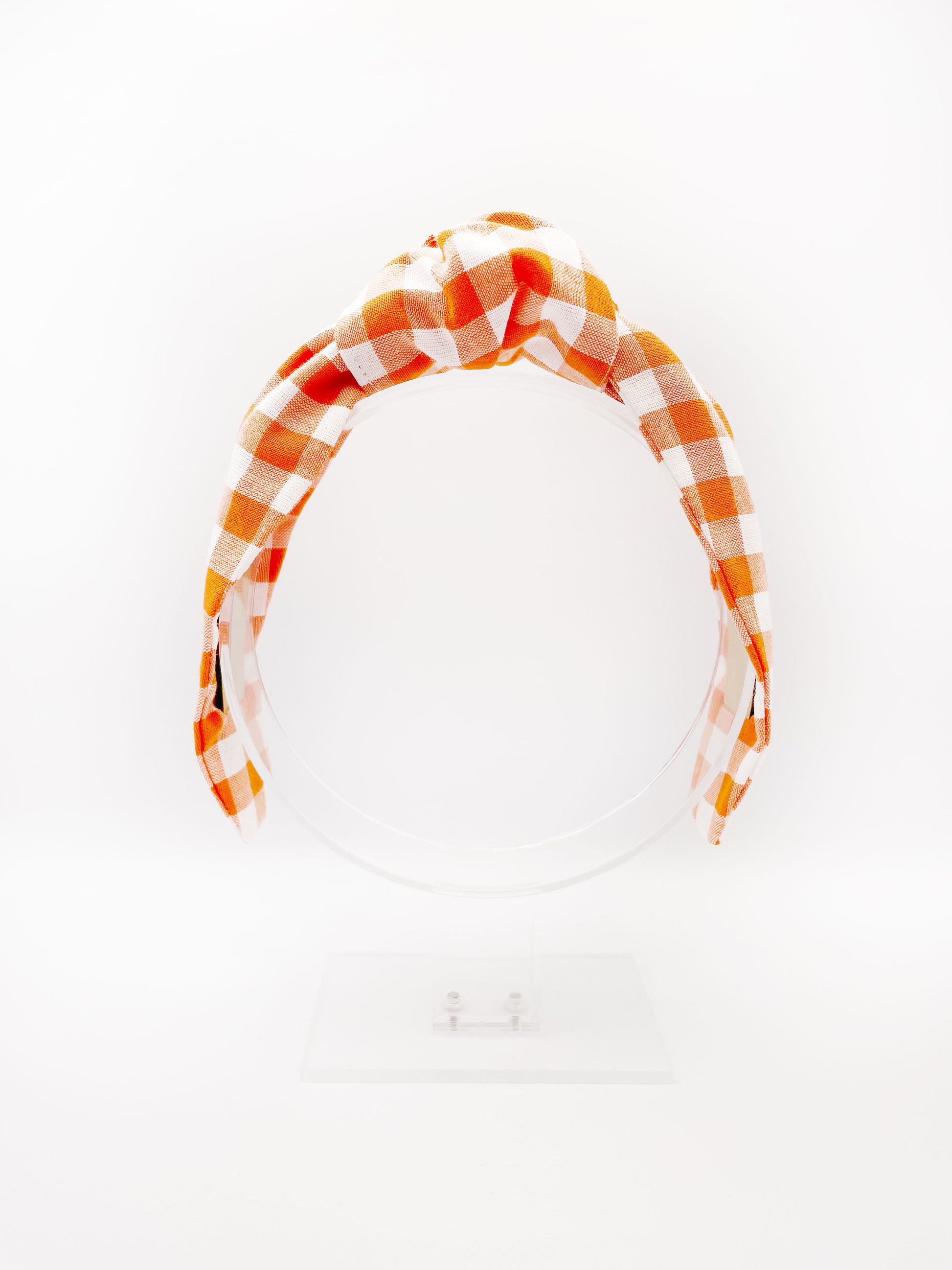 A knotted headband with an orange and white gingham plaid print. The headband is displayed on an acrylic headband display.