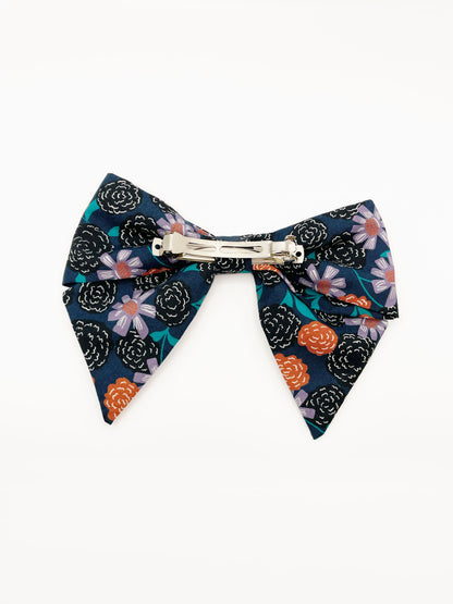 A large handmade hair bow with vivid orange, lavender, and black flowers, accented with fresh green leaves. The cotton fabric is attached to a sturdy barrette clip.