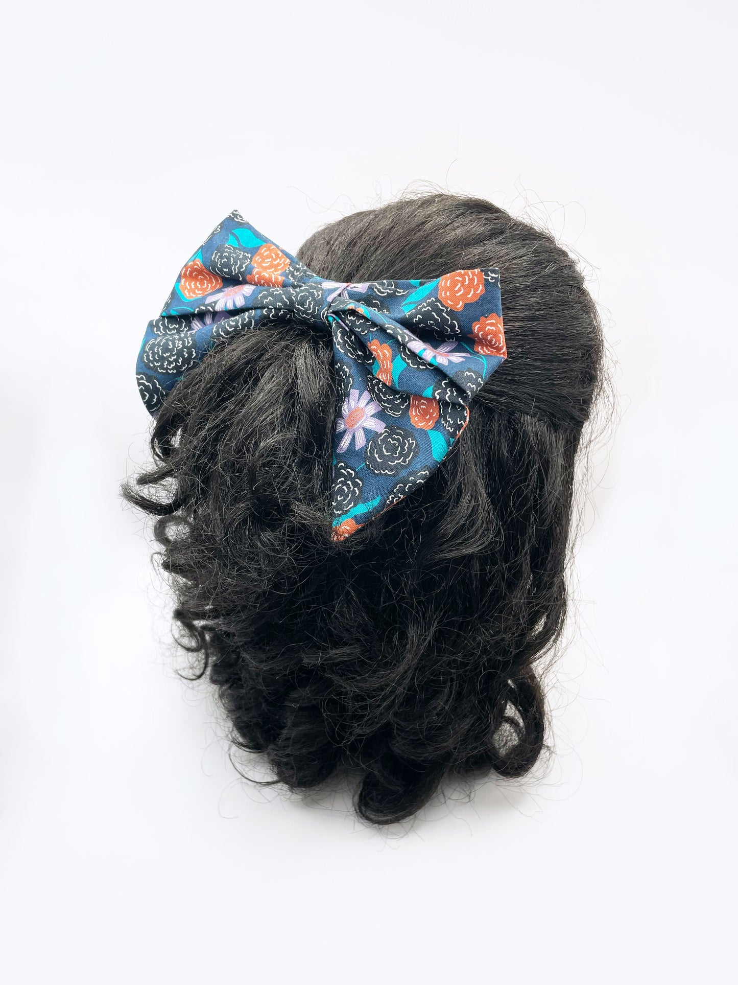A large handmade hair bow with vivid orange, lavender, and black flowers, accented with fresh green leaves. The bow is modeled on a mannequin head.
