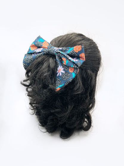 A large handmade hair bow with vivid orange, lavender, and black flowers, accented with fresh green leaves. The bow is modeled on a mannequin head.