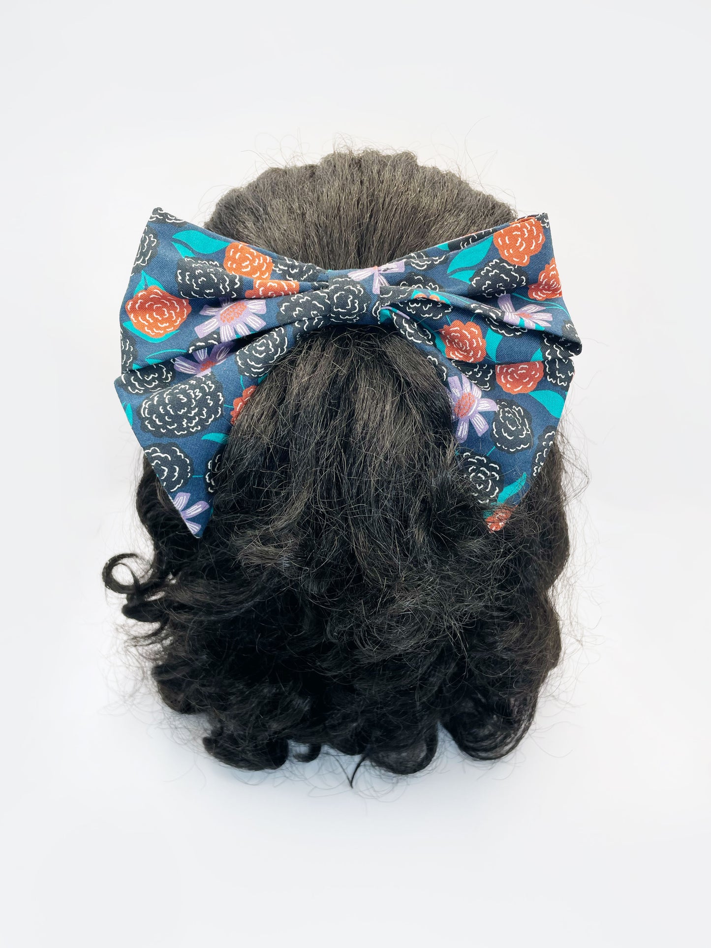 A large handmade hair bow with vivid orange, lavender, and black flowers, accented with fresh green leaves. The bow is modeled on a mannequin head.