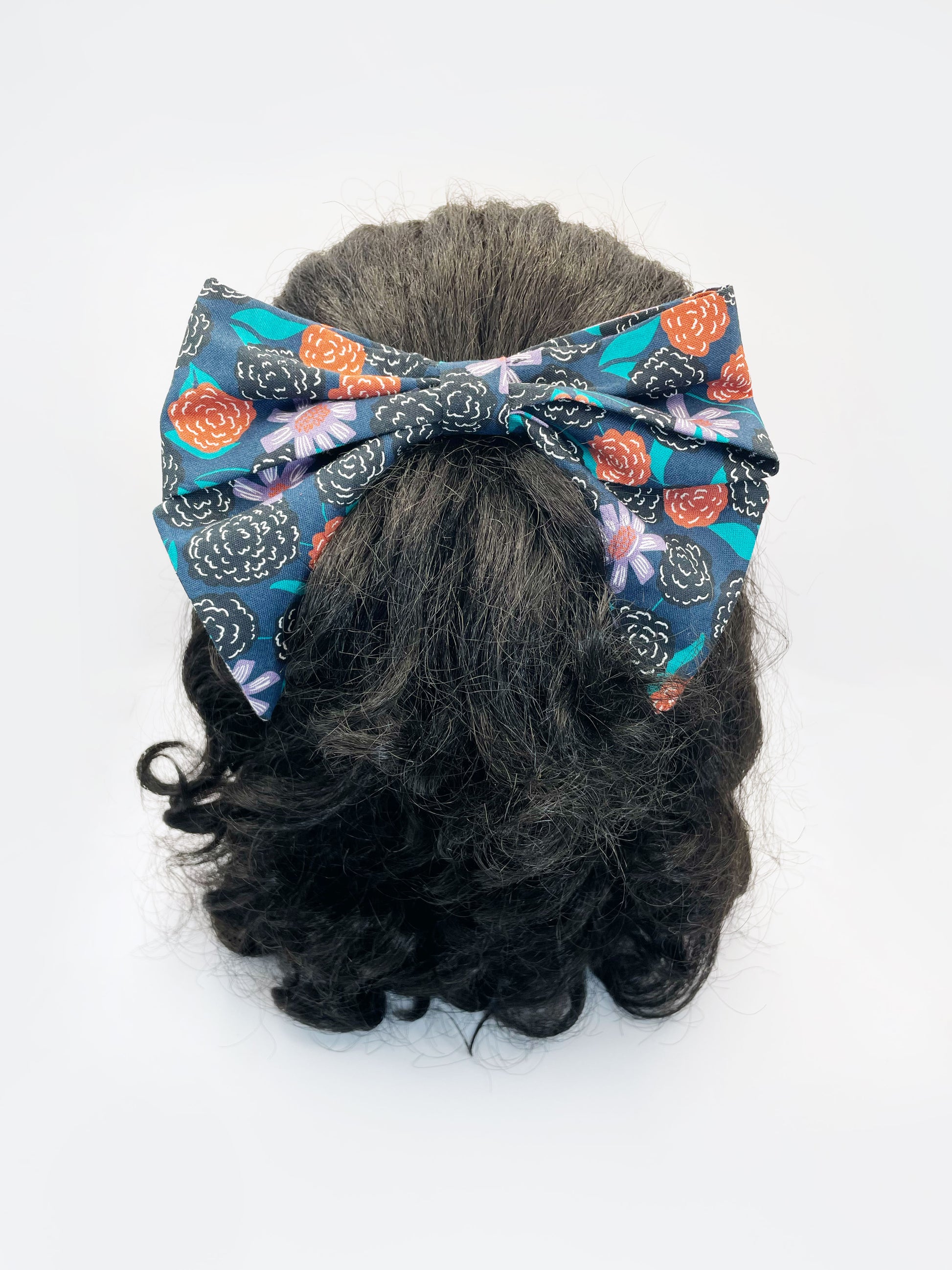 A large handmade hair bow with vivid orange, lavender, and black flowers, accented with fresh green leaves. The bow is modeled on a mannequin head.