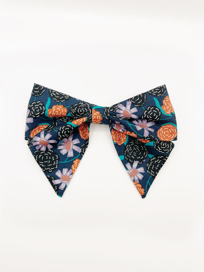 A large handmade hair bow with vivid orange, lavender, and black flowers, accented with fresh green leaves.