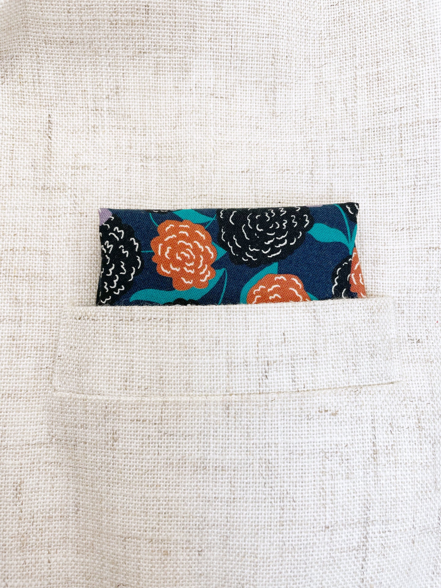 A navy blue pocket square with vivid orange, lavender, and black flowers, accented with fresh green leaves.
