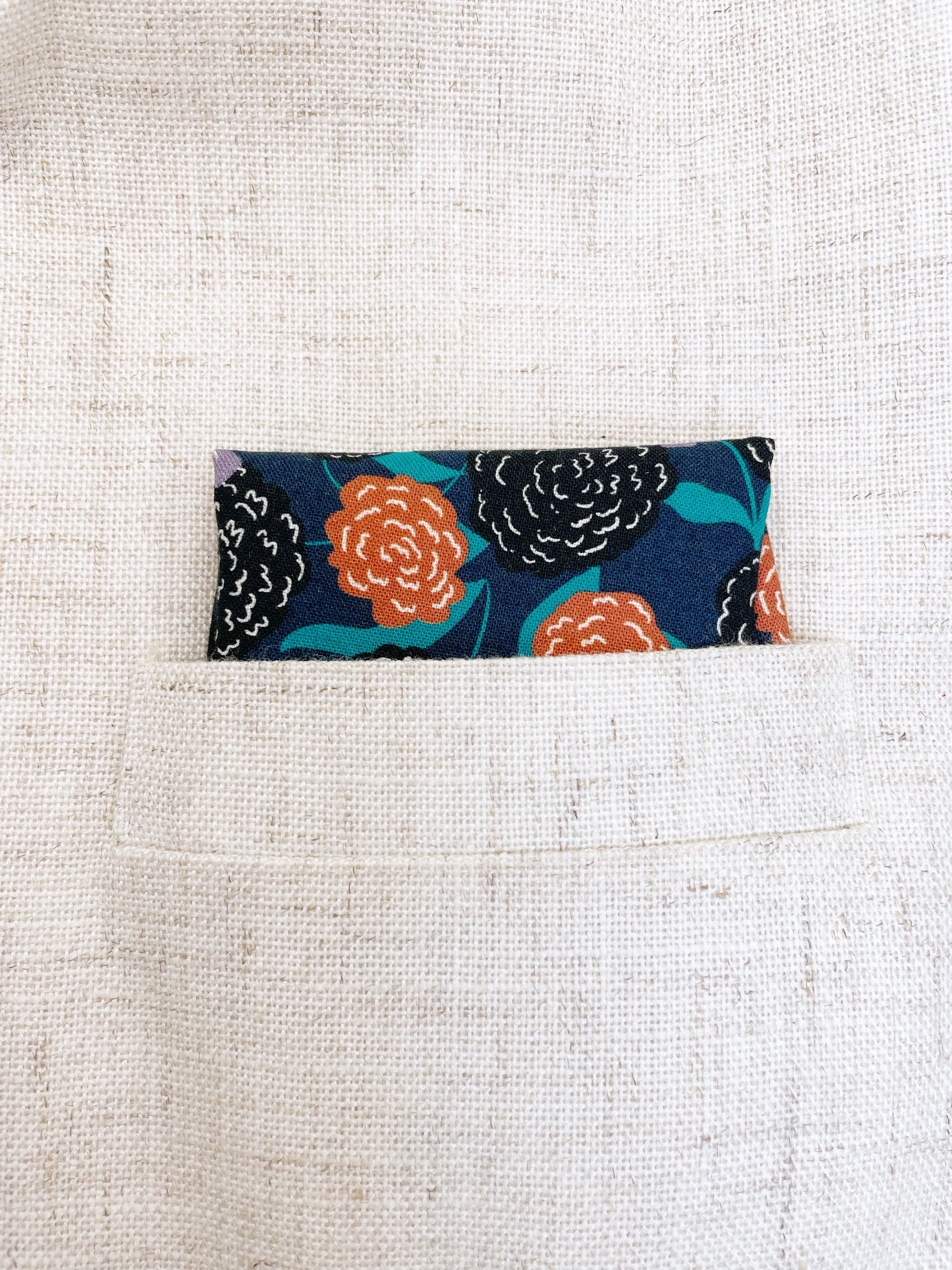 A navy blue pocket square with vivid orange, lavender, and black flowers, accented with fresh green leaves.