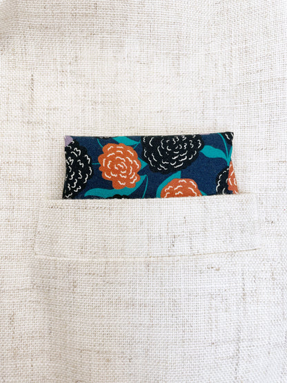 A navy blue pocket square with vivid orange, lavender, and black flowers, accented with fresh green leaves.