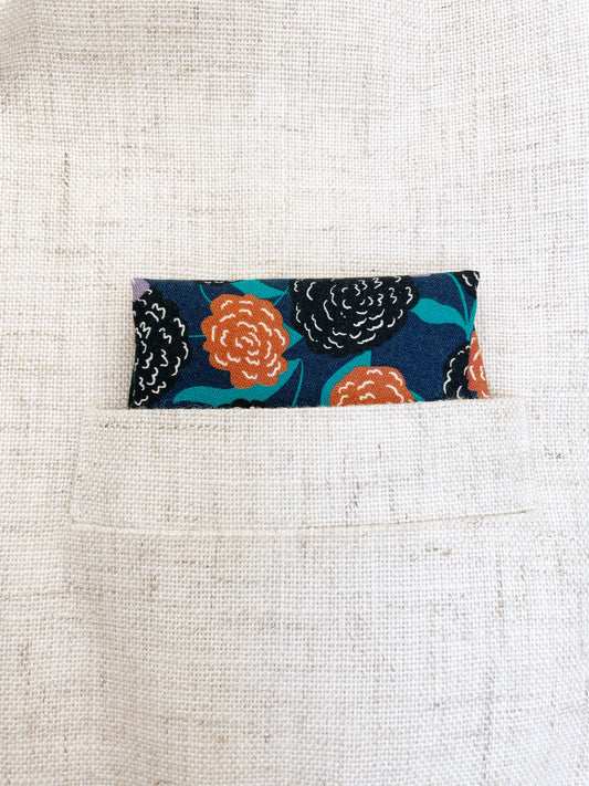 A navy blue pocket square with vivid orange, lavender, and black flowers, accented with fresh green leaves.
