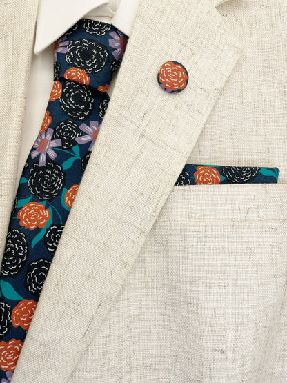 A navy blue pocket square with vivid orange, lavender, and black flowers, accented with fresh green leaves. The pocket square is shown with a matching necktie and lapel pin, along with a neutral suit jacket.