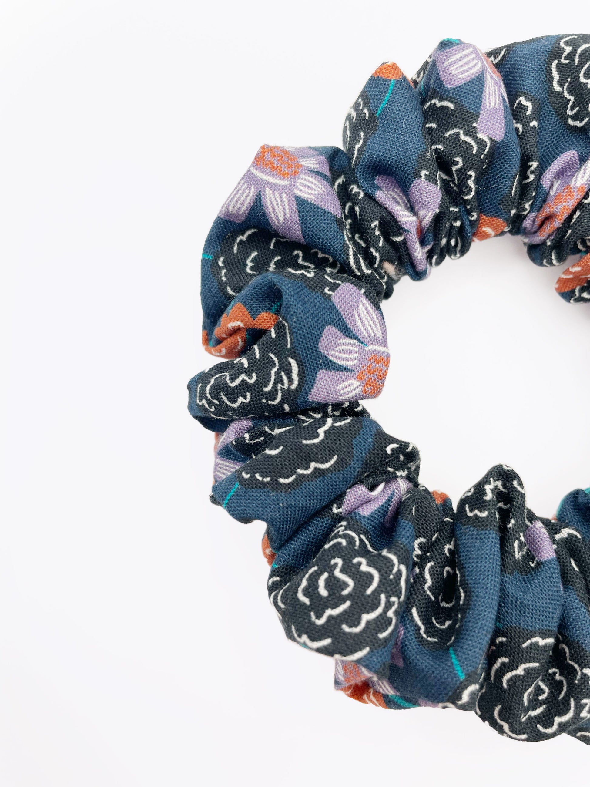 A scrunchie with vivid orange, lavender, and black flowers, accented with fresh green leaves.