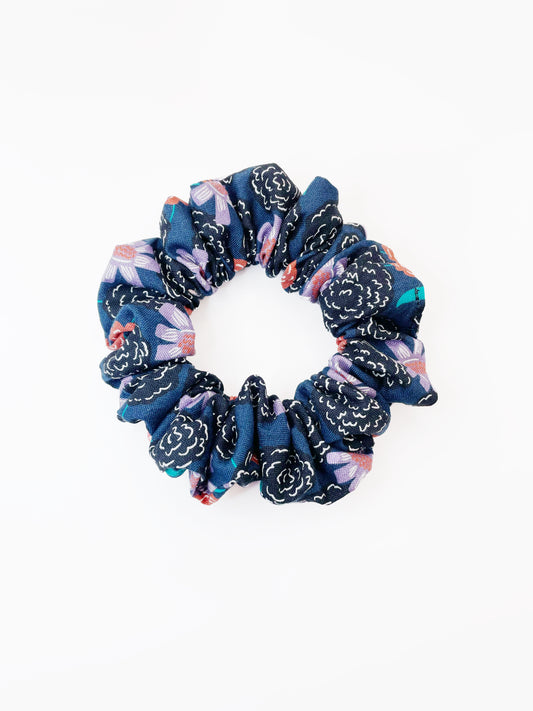 A scrunchie with vivid orange, lavender, and black flowers, accented with fresh green leaves.