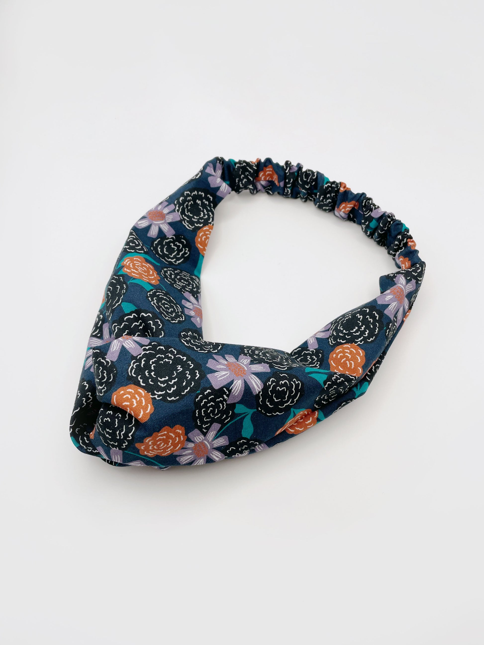 A twist headband with vivid orange, lavender, and black flowers, accented with fresh green leaves. The elastic band in the back provides support without slipping. 