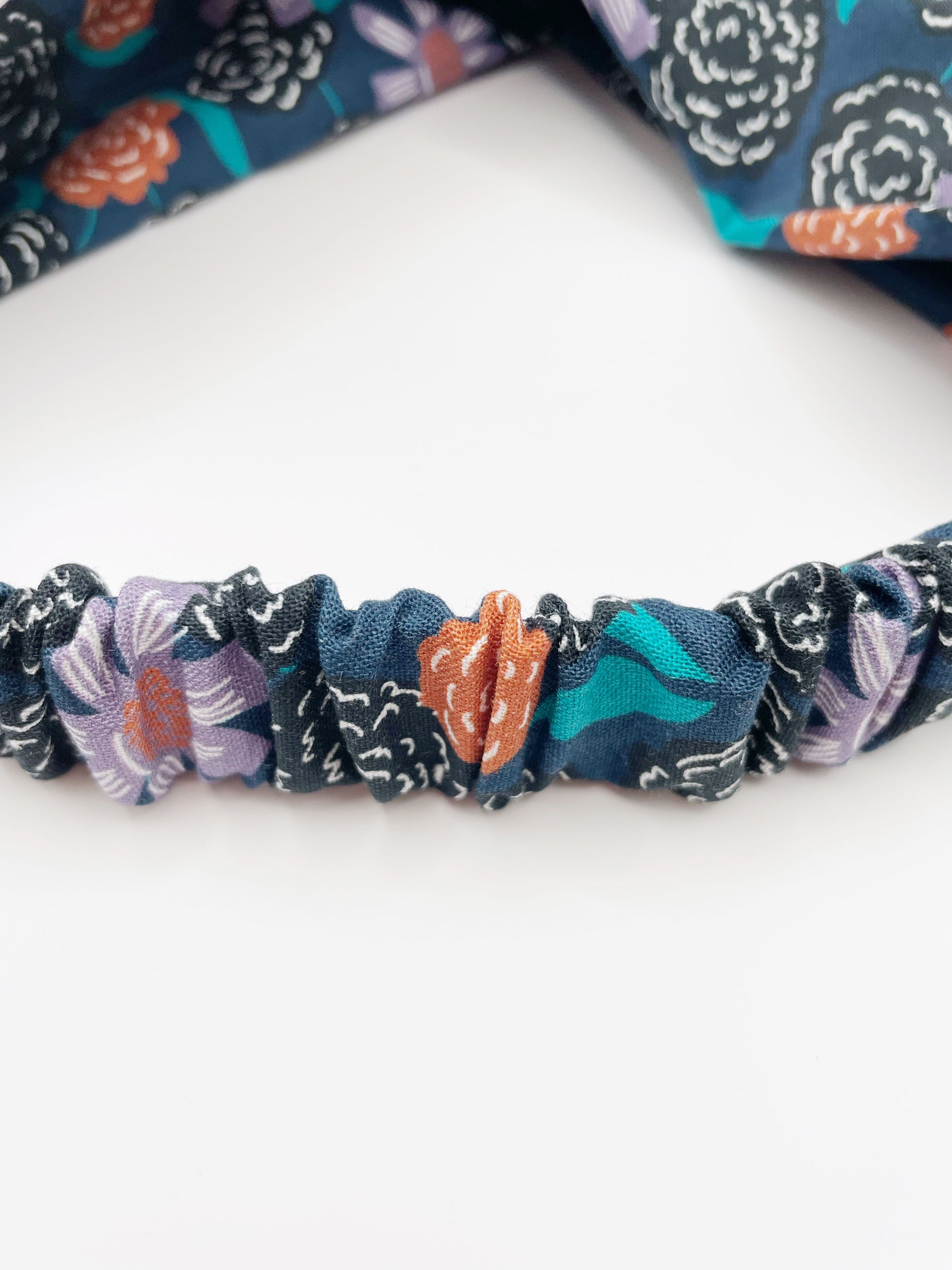 A twist headband with vivid orange, lavender, and black flowers, accented with fresh green leaves. The elastic band in the back provides support without slipping. 