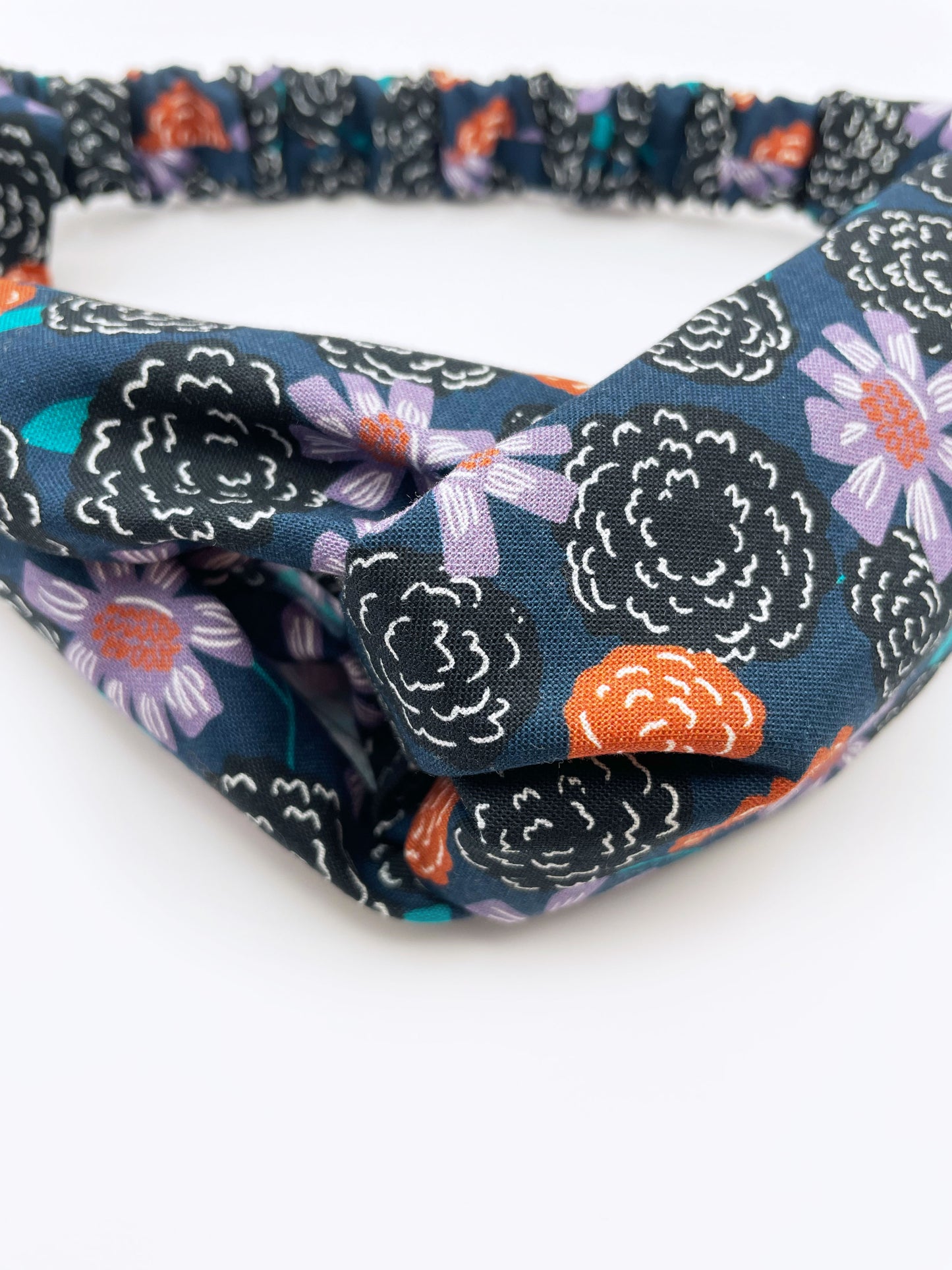 A twist headband with vivid orange, lavender, and black flowers, accented with fresh green leaves. The elastic band in the back provides support without slipping. 