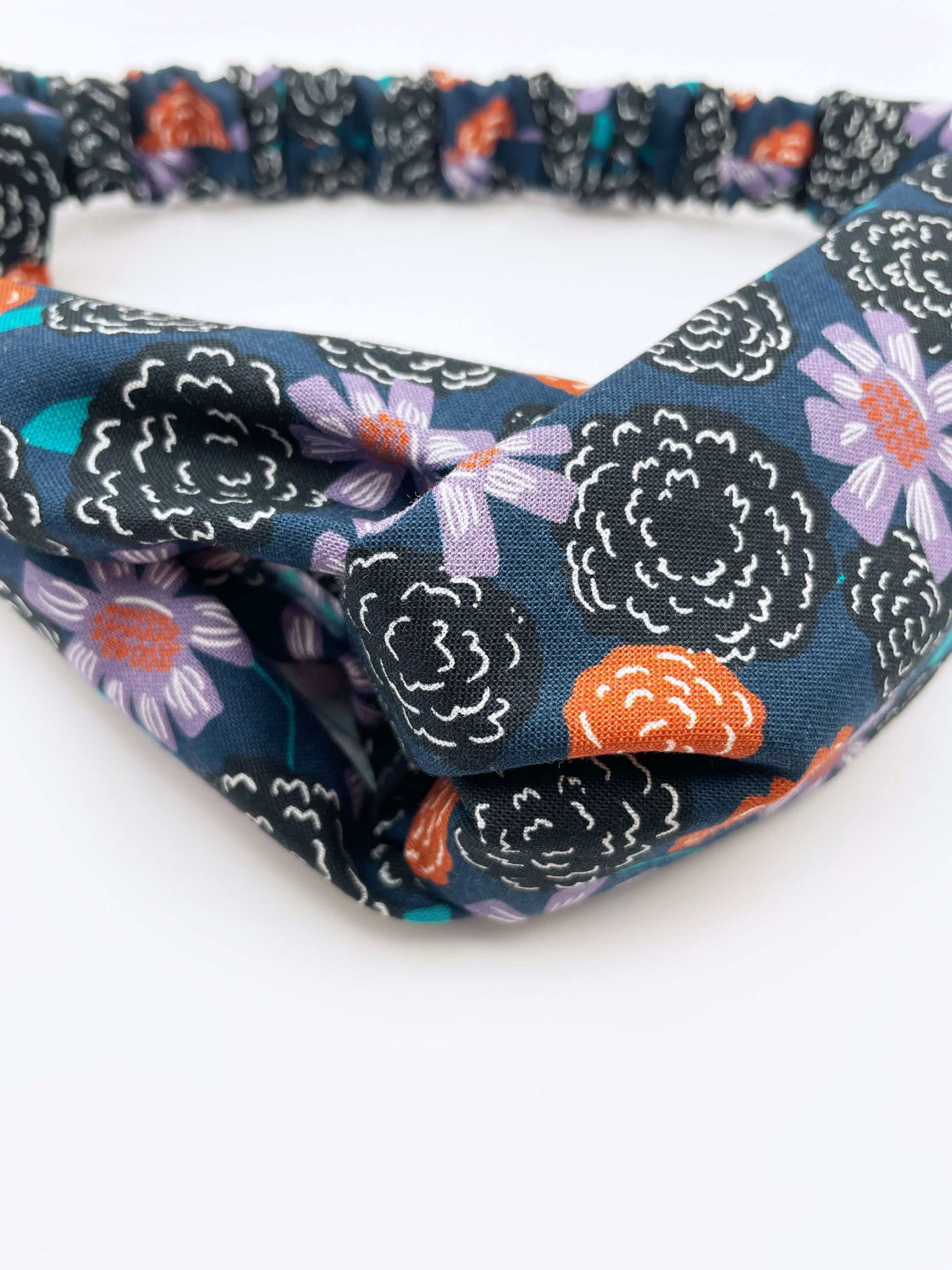 A twist headband with vivid orange, lavender, and black flowers, accented with fresh green leaves. The elastic band in the back provides support without slipping. 