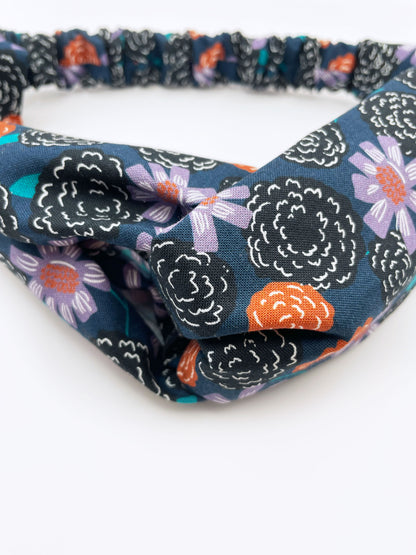 A twist headband with vivid orange, lavender, and black flowers, accented with fresh green leaves. The elastic band in the back provides support without slipping. 