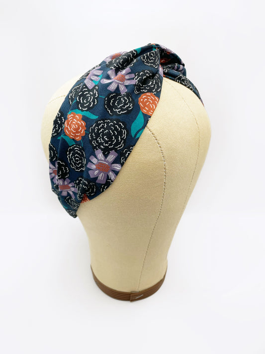 A twist headband with vivid orange, lavender, and black flowers, accented with fresh green leaves. The headband is modeled on a mannequin head.