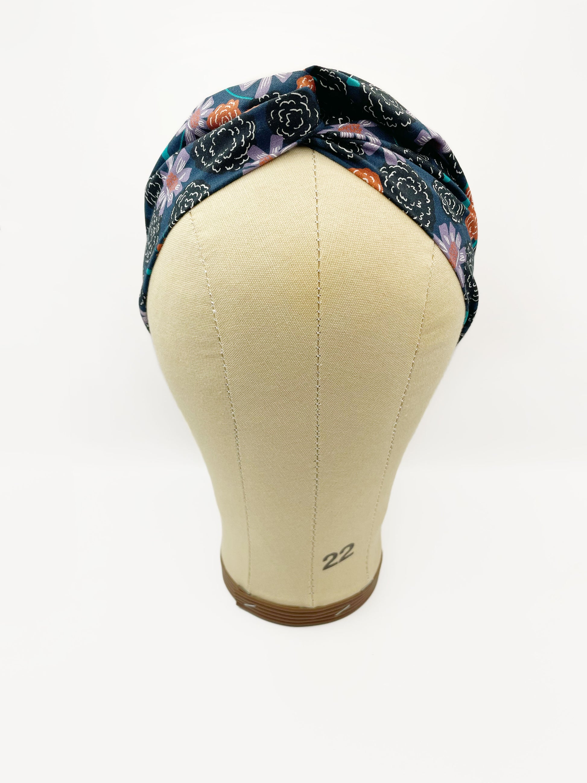 A twist headband with vivid orange, lavender, and black flowers, accented with fresh green leaves. The headband is modeled on a mannequin head.