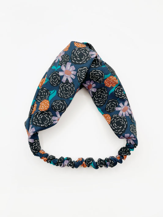 A twist headband with vivid orange, lavender, and black flowers, accented with fresh green leaves. The elastic band in the back provides support without slipping. 