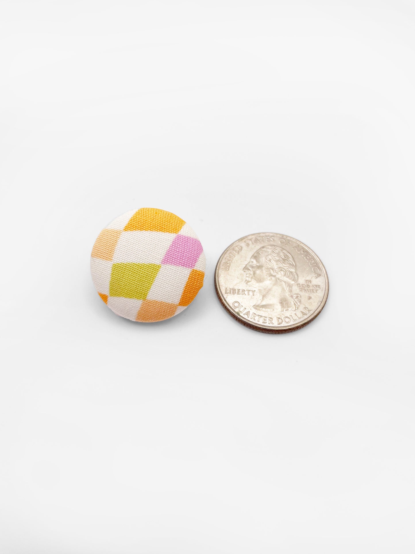 A fabric-covered lapel pin with a pastel checkered print. The pin is next to a quarter for size reference.