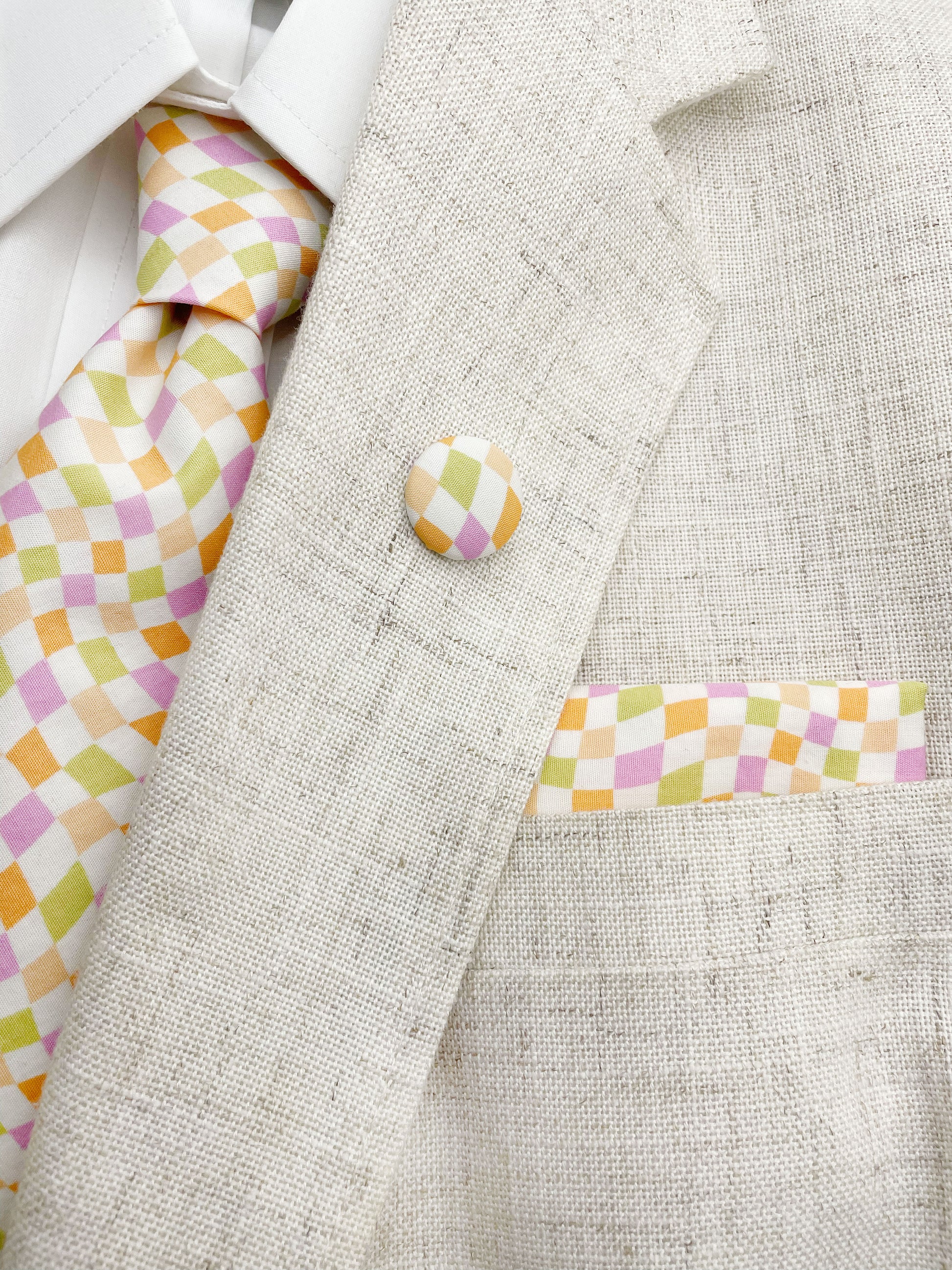 A handmade necktie with a pastel checkered print shown with a matching pocket square and lapel pin.