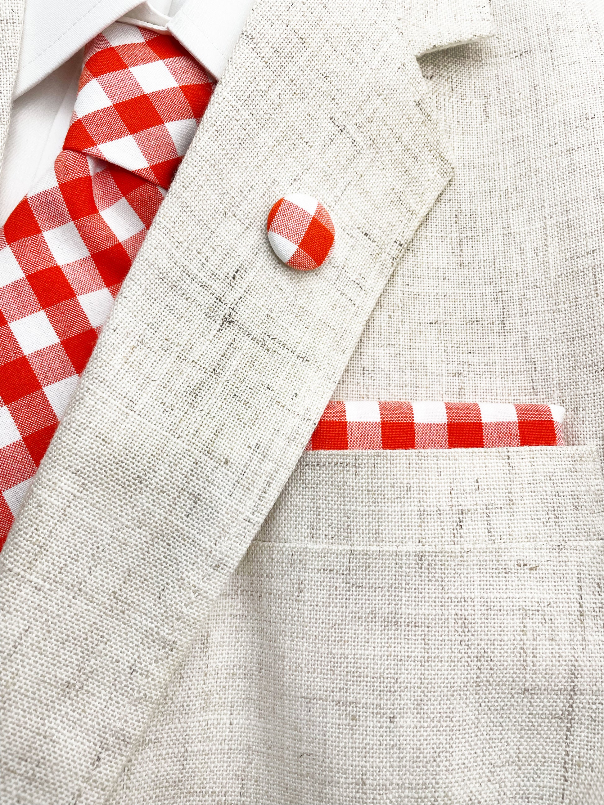 A cotton necktie with a vibrant red gingham plaid print shown with a matching pocket square and lapel pin.