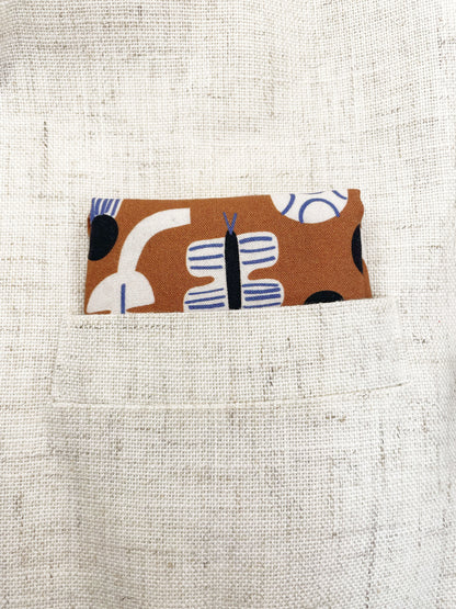 A pocket square with an abstract print featuring flowers, leaves, mushrooms, and butterflies on a rich terracotta background. The color palette also consists of cream, black, and blue.