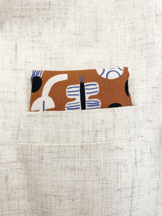 A pocket square with an abstract print featuring flowers, leaves, mushrooms, and butterflies on a rich terracotta background. The color palette also consists of cream, black, and blue.