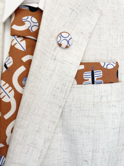 A pocket square with an abstract print featuring flowers, leaves, mushrooms, and butterflies on a rich terracotta background. The color palette also consists of cream, black, and blue. The pocket square is shown with a matching necktie and lapel pin, along with a neutral suit jacket.