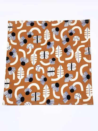A pocket square with an abstract print featuring flowers, leaves, mushrooms, and butterflies on a rich terracotta background. The color palette also consists of cream, black, and blue.