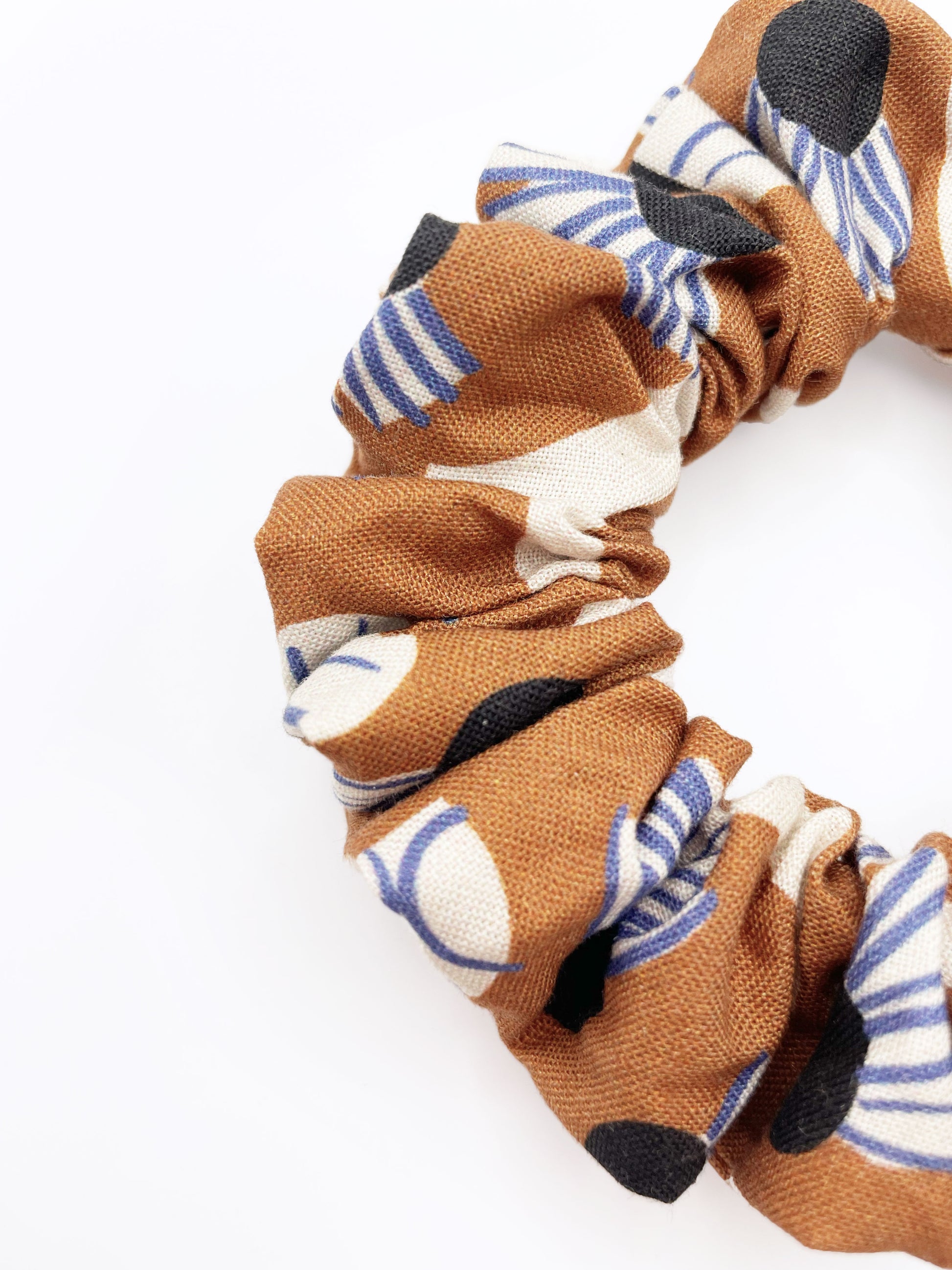 A scrunchie with an abstract print featuring flowers, leaves, and butterflies on a rich terracotta background. The color palette also consists of cream, black, and blue.
