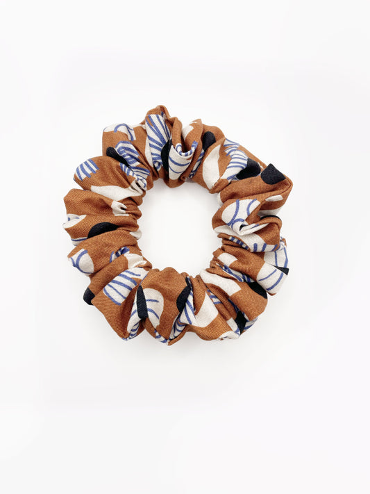 A scrunchie with an abstract print featuring flowers, leaves, and butterflies on a rich terracotta background. The color palette also consists of cream, black, and blue.