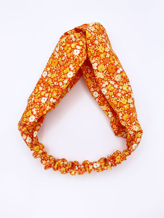 An orange cotton twist headband with a colorful ditsy floral print. The headband has an elastic band in the back to provide comfort without slipping.