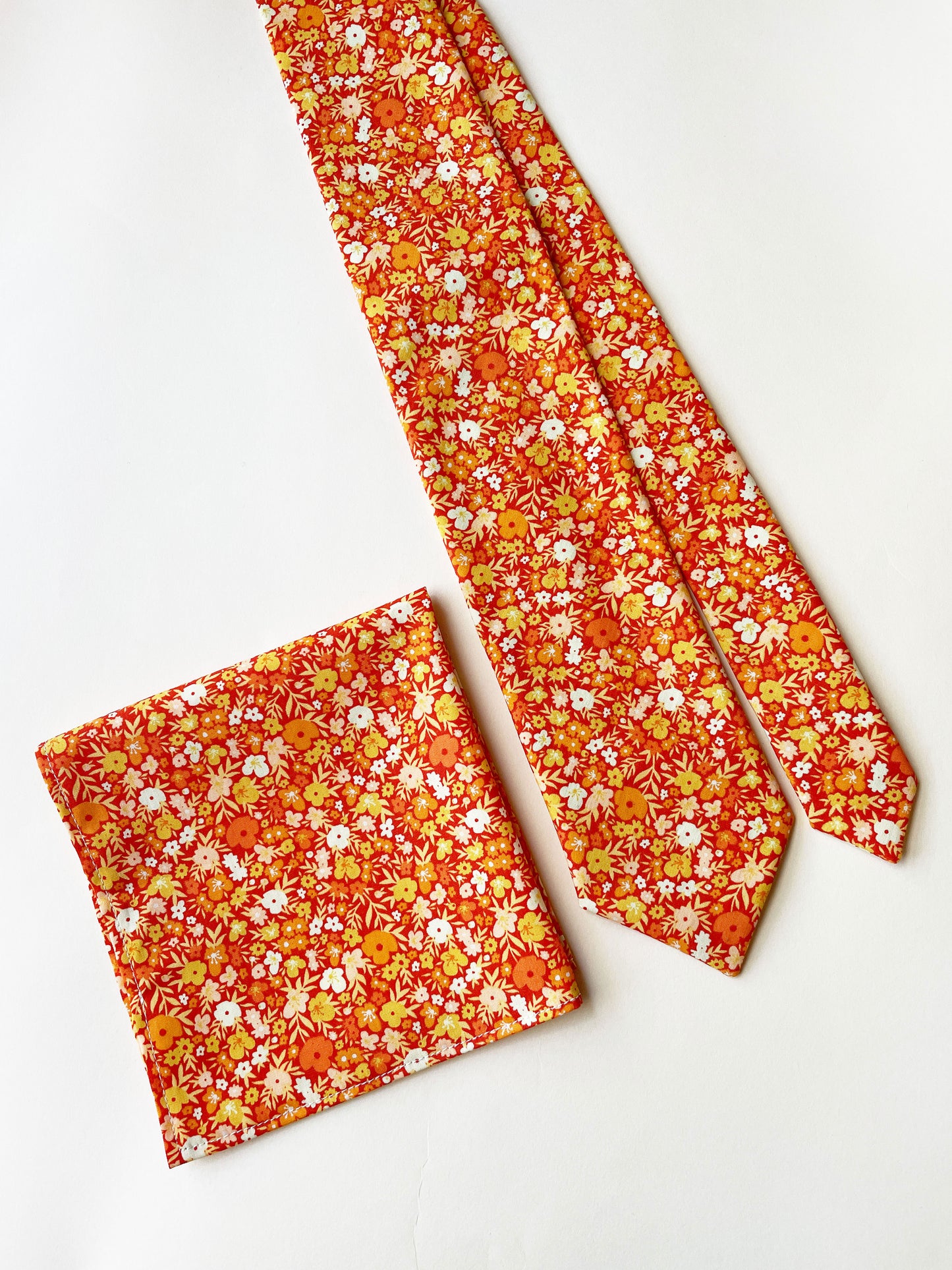 A handmade pocket square featuring vibrant orange and yellow flowers next to a matching necktie.