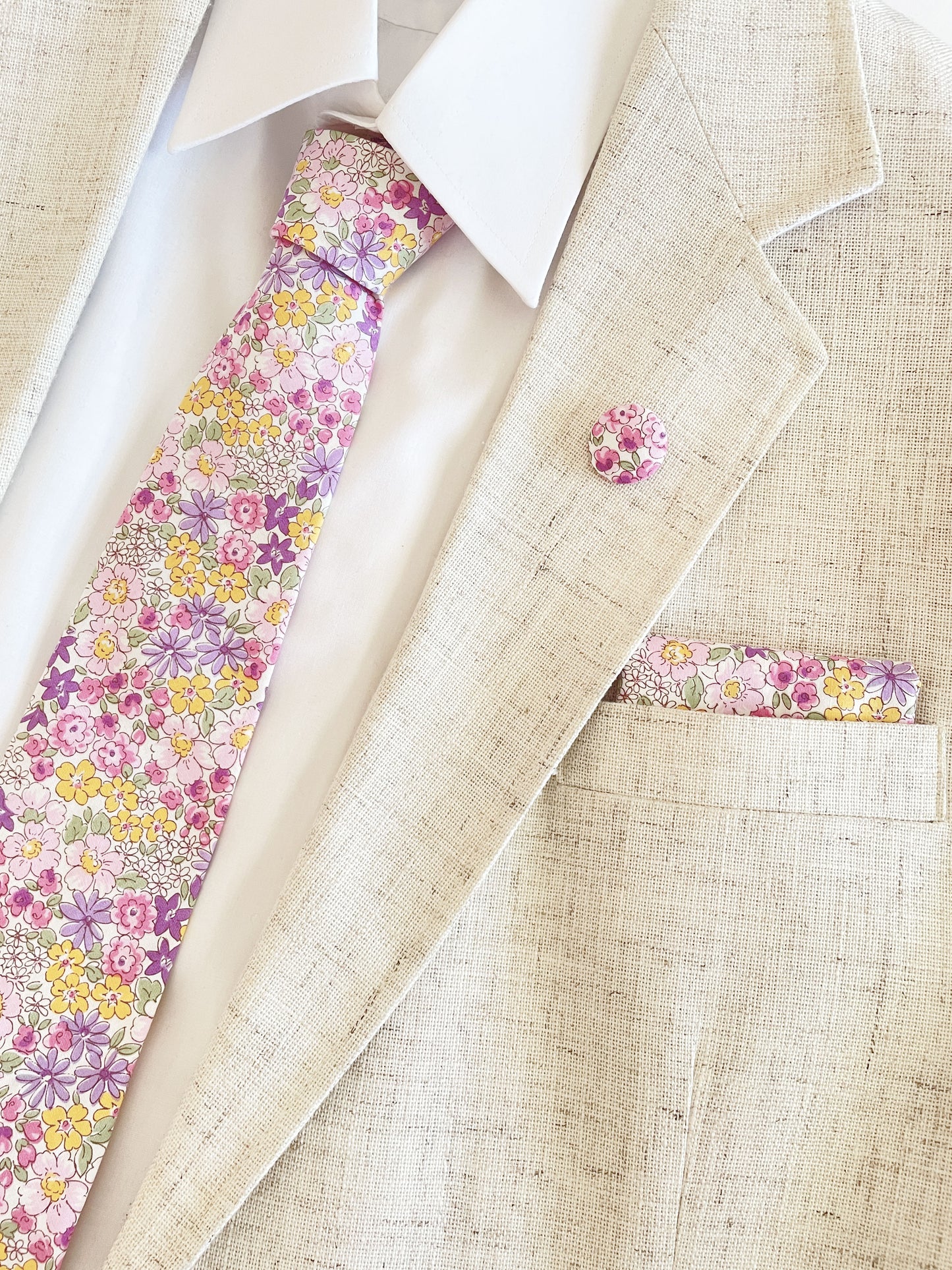 A handmade purple floral necktie paired with a matching pocket square and lapel pin, shown with a white button down shirt in a beige suit jacket.