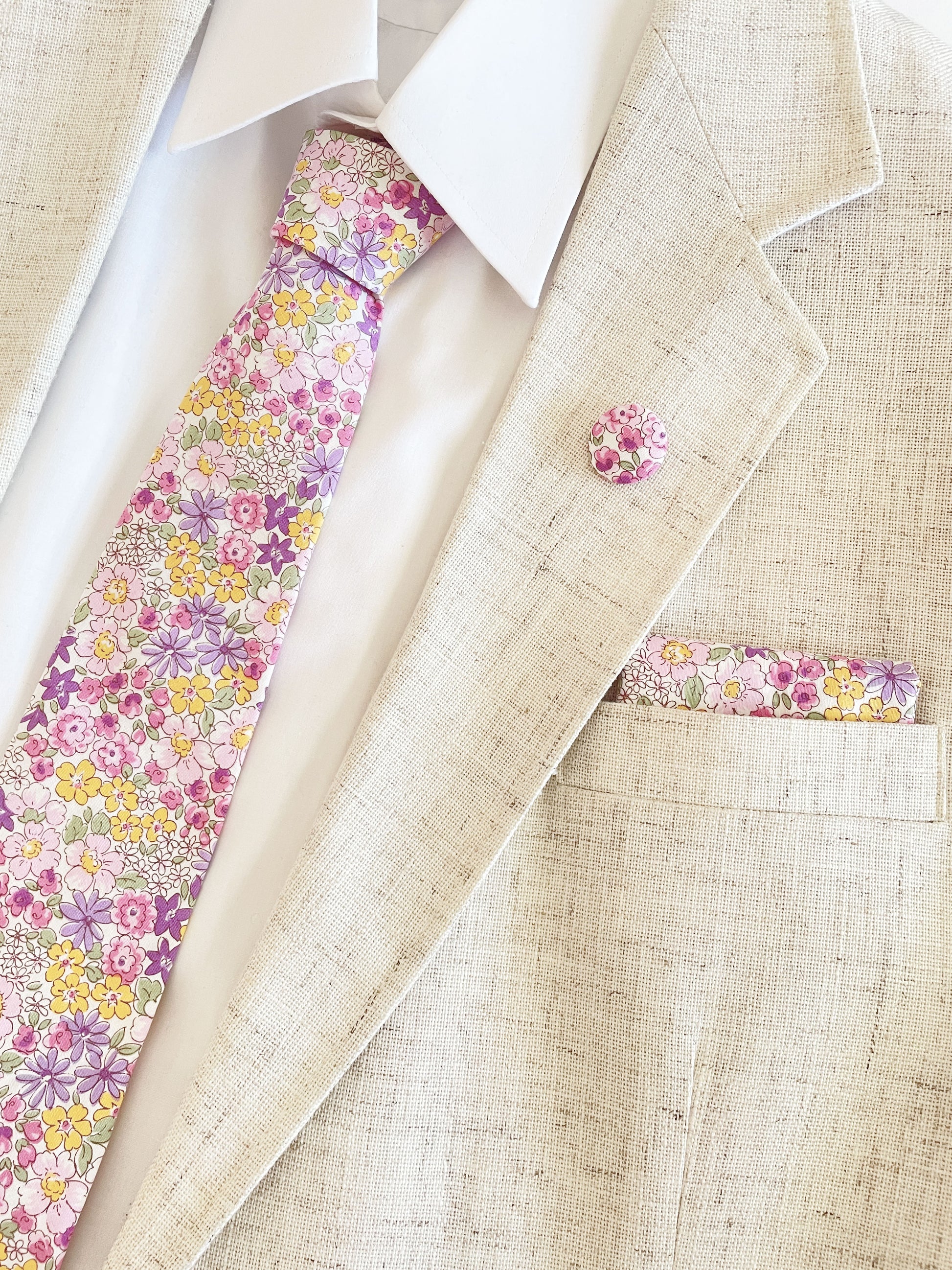 A handmade purple floral necktie paired with a matching pocket square and lapel pin, shown with a white button down shirt in a beige suit jacket.