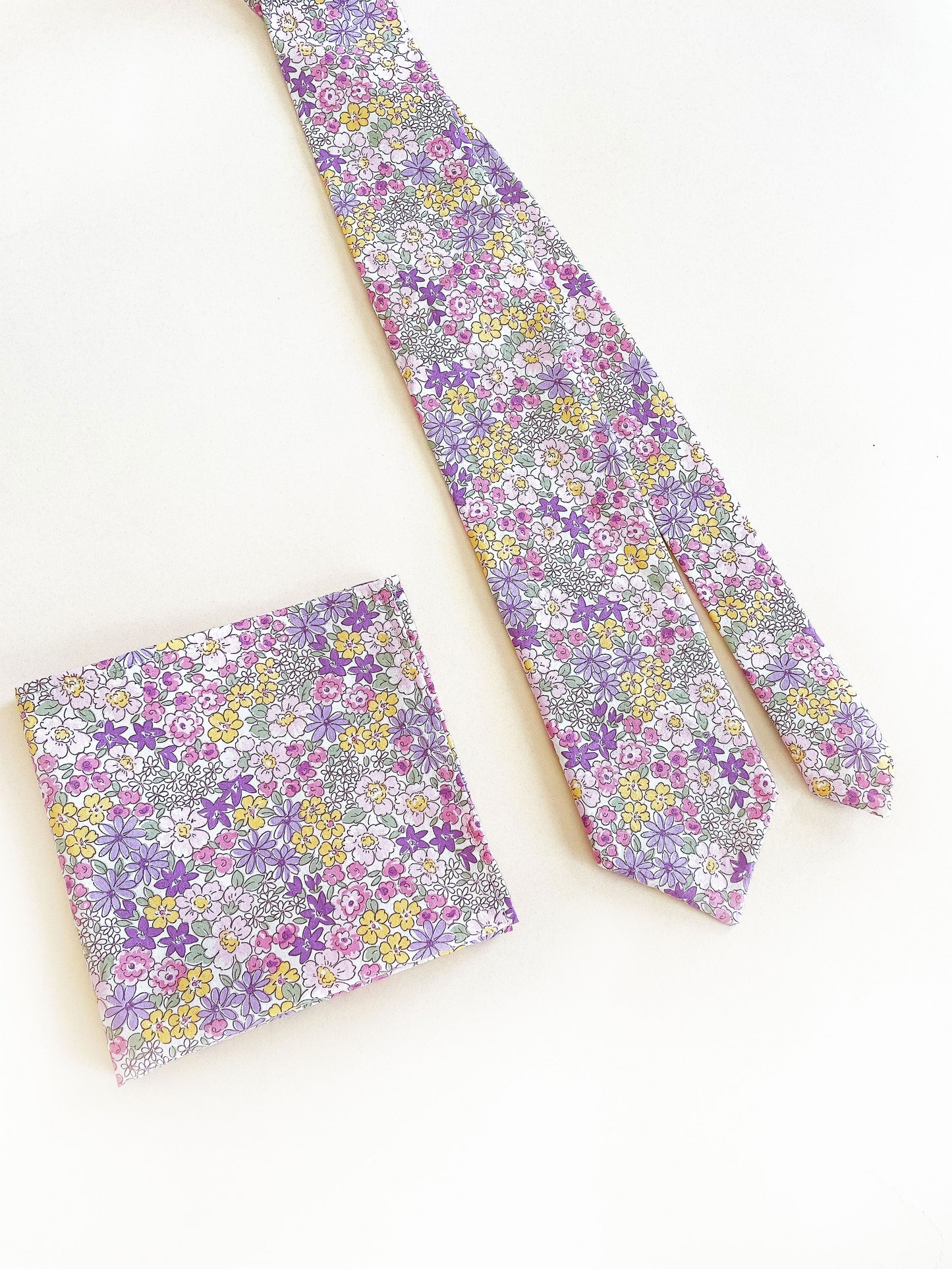 A handmade pocket square with purple flowers with a matching necktie against an off white background.