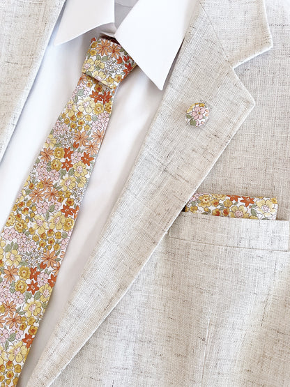 A handmade yellow floral necktie paired with a matching pocket square and lapel pin, shown with a white button down shirt in a beige suit jacket.