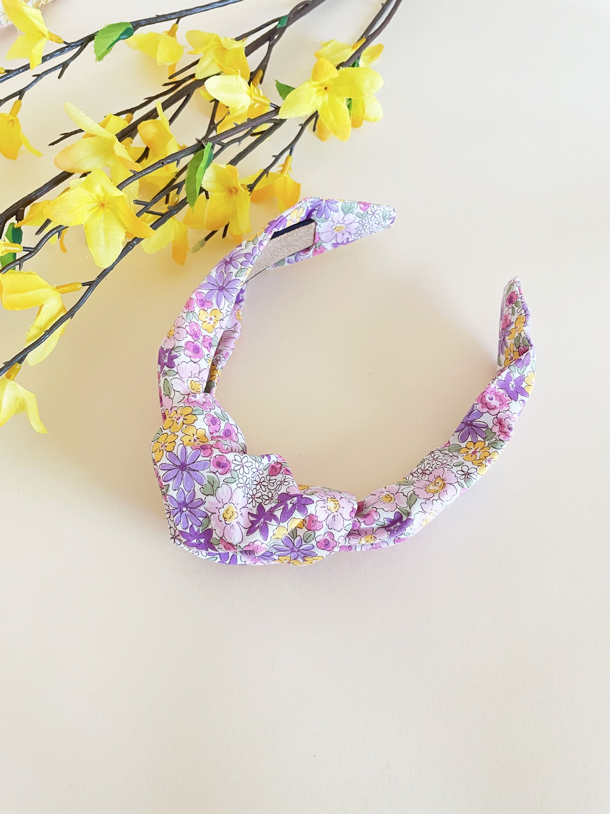 Handmade knotted headband with purple flowers against an off white background next to yellow flowers..